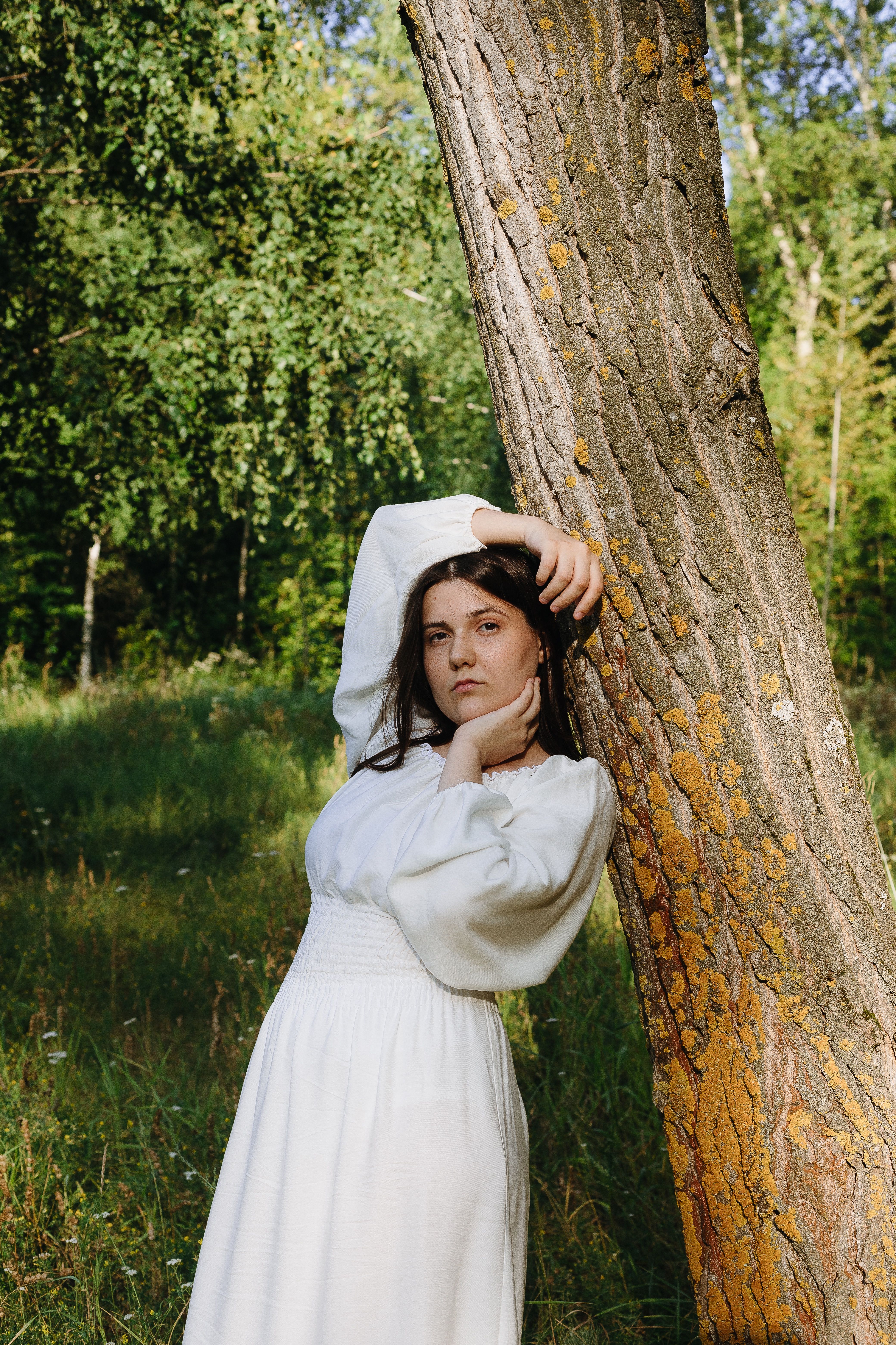 Woman in a white dress posing by a tree in a sunny forest.