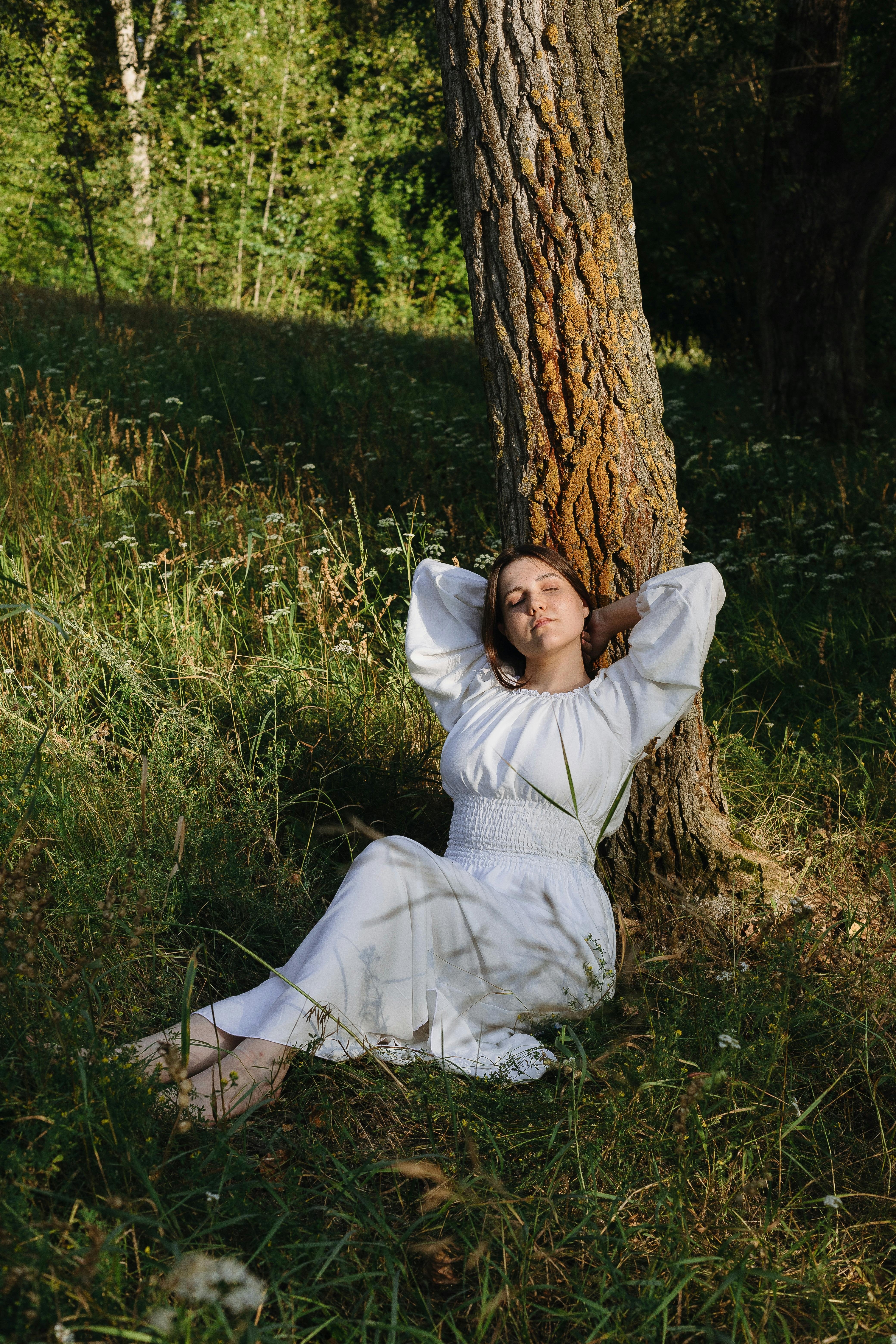Free Young woman in white dress resting against tree in a sunlit forest. Stock Photo