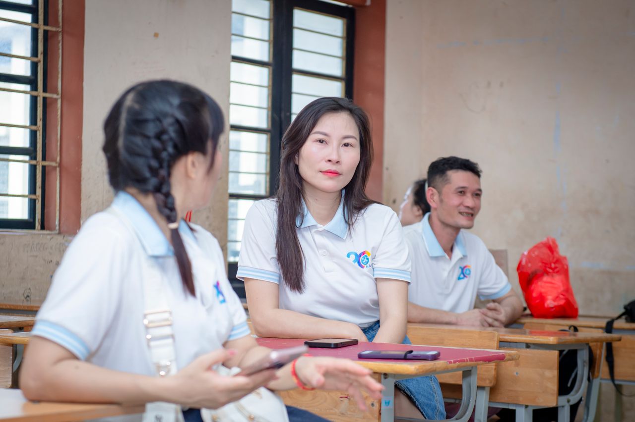 Three adults having a casual discussion in a classroom setting in Hanoi, Vietnam.