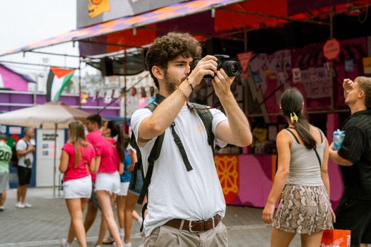 A young man takes photos at an outdoor urban festival surrounded by vibrant stalls and people.