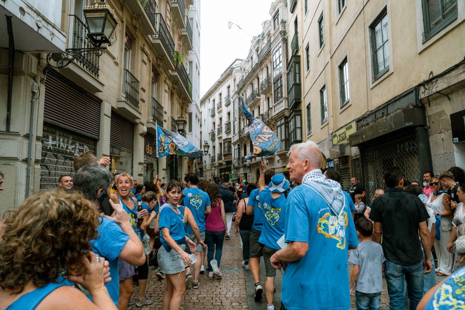 Festival callejero colorido con gente vestida de azul, banderas festivas y arquitectura europea clásica.