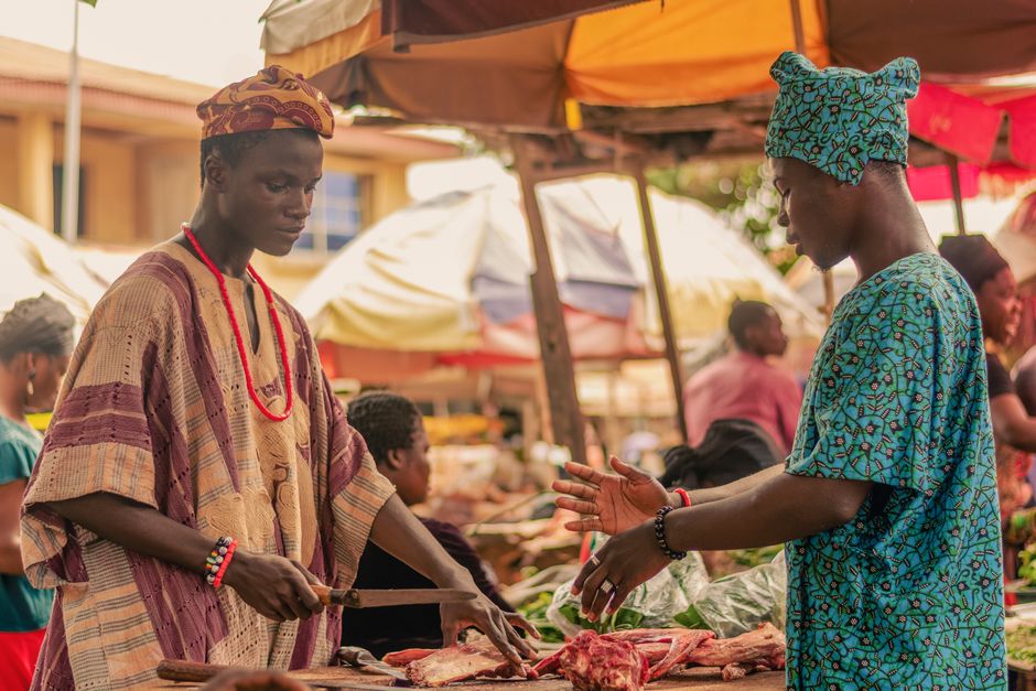 Dark-skinned Nigerian woman in a vibrant Ankara blouse smiling as she reviews a tablet showing sales figures at her Lagos food market stall, fresh tomatoes and peppers arranged on the table in front of her, bright natural daylight, photorealistic