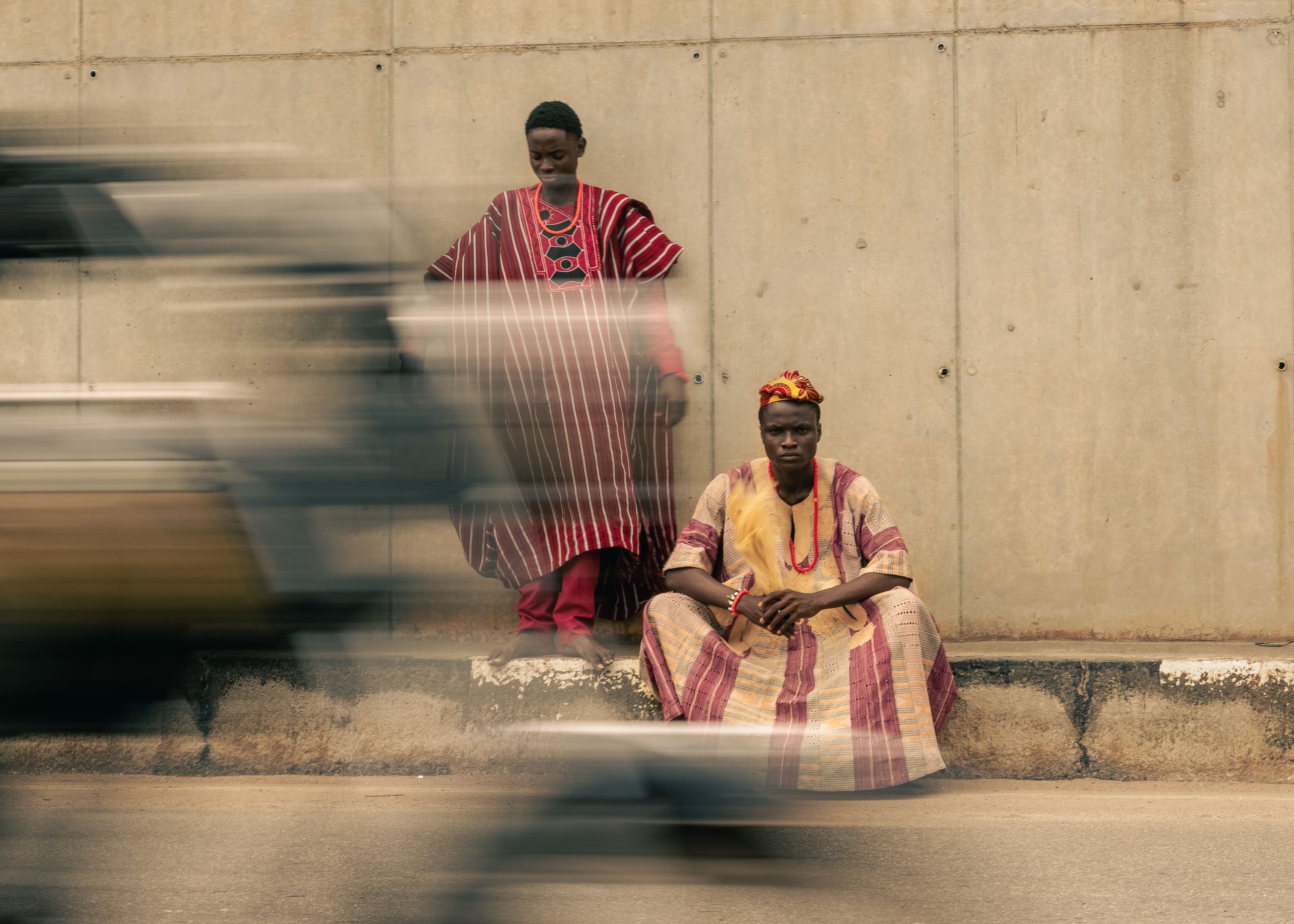 Two men in traditional African attire seated by a roadside, blurred motion of traffic.