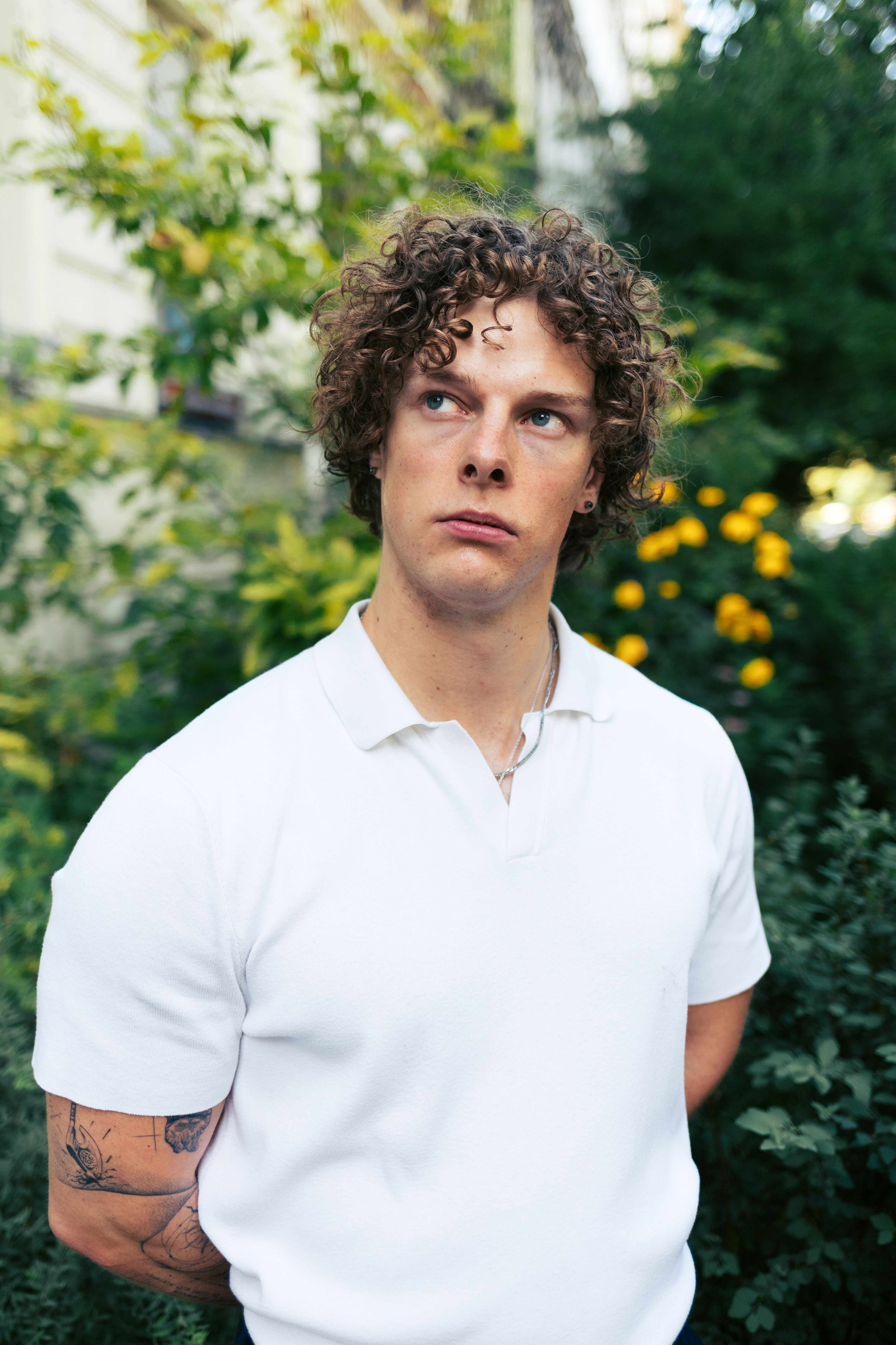 Portrait of a young adult man with curly hair in a white shirt, against a leafy background in Berlin.