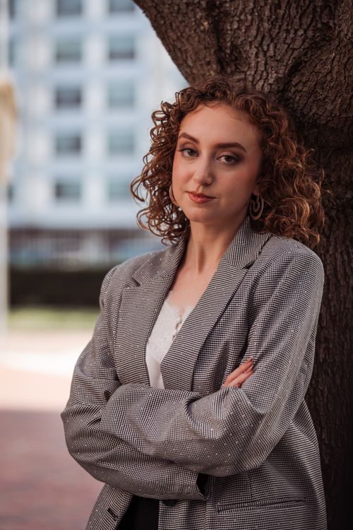 Free Young woman in a gray blazer leans against a tree in an urban setting, exuding confidence. Stock Photo
