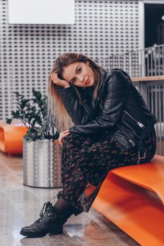 Fashionable woman in leather jacket sitting on an orange bench indoors, exuding casual elegance.