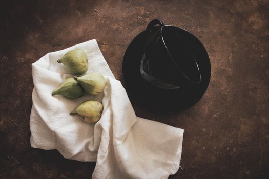 An artistic arrangement of green figs on a cloth next to a black cup on a rustic background.