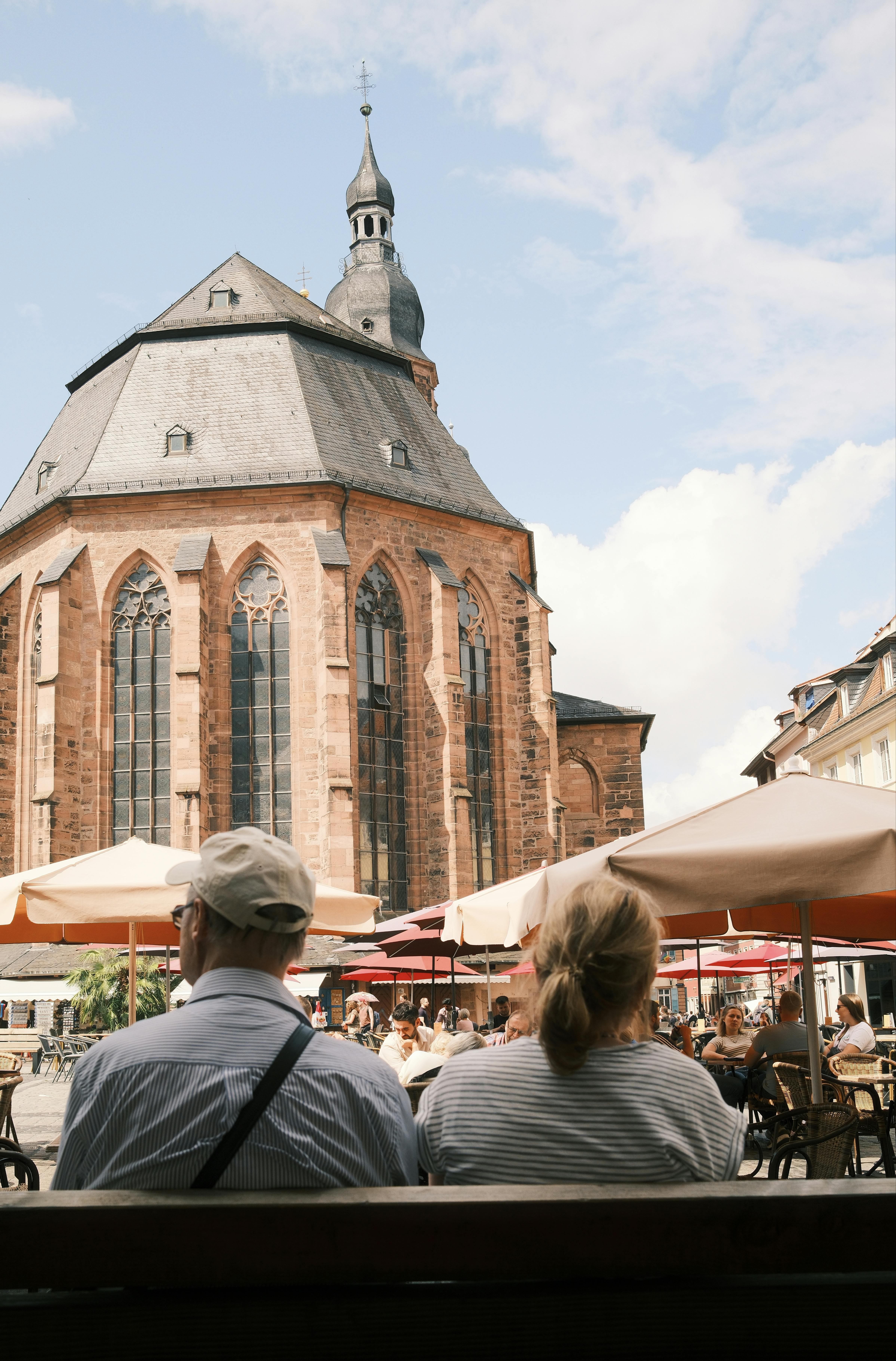 Senior couple enjoys a sunny day at an outdoor cafe beside a historic church in Heidelberg.