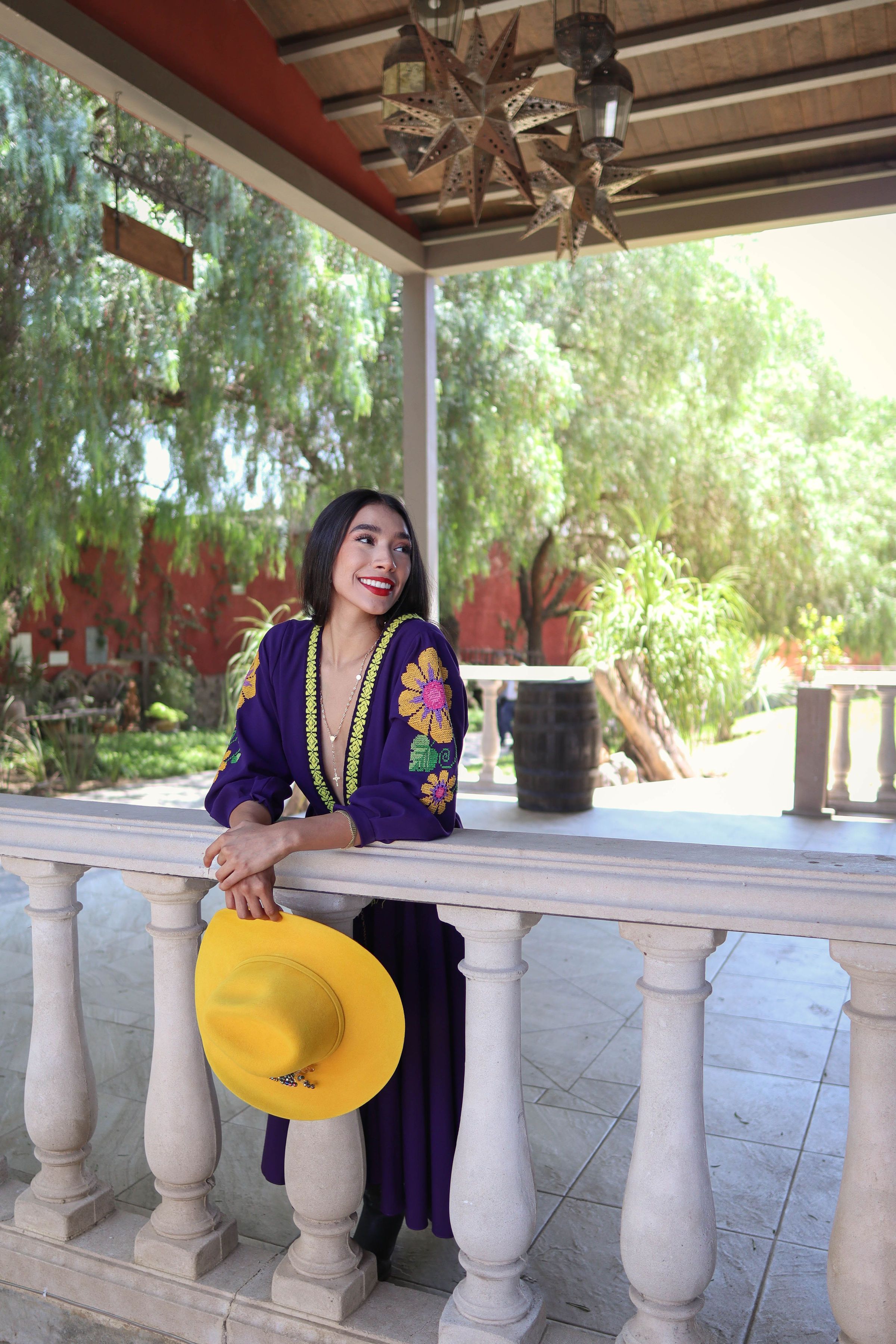 Charming woman in purple traditional Mexican dress, holding a yellow hat, enjoying a sunny day outdoors.