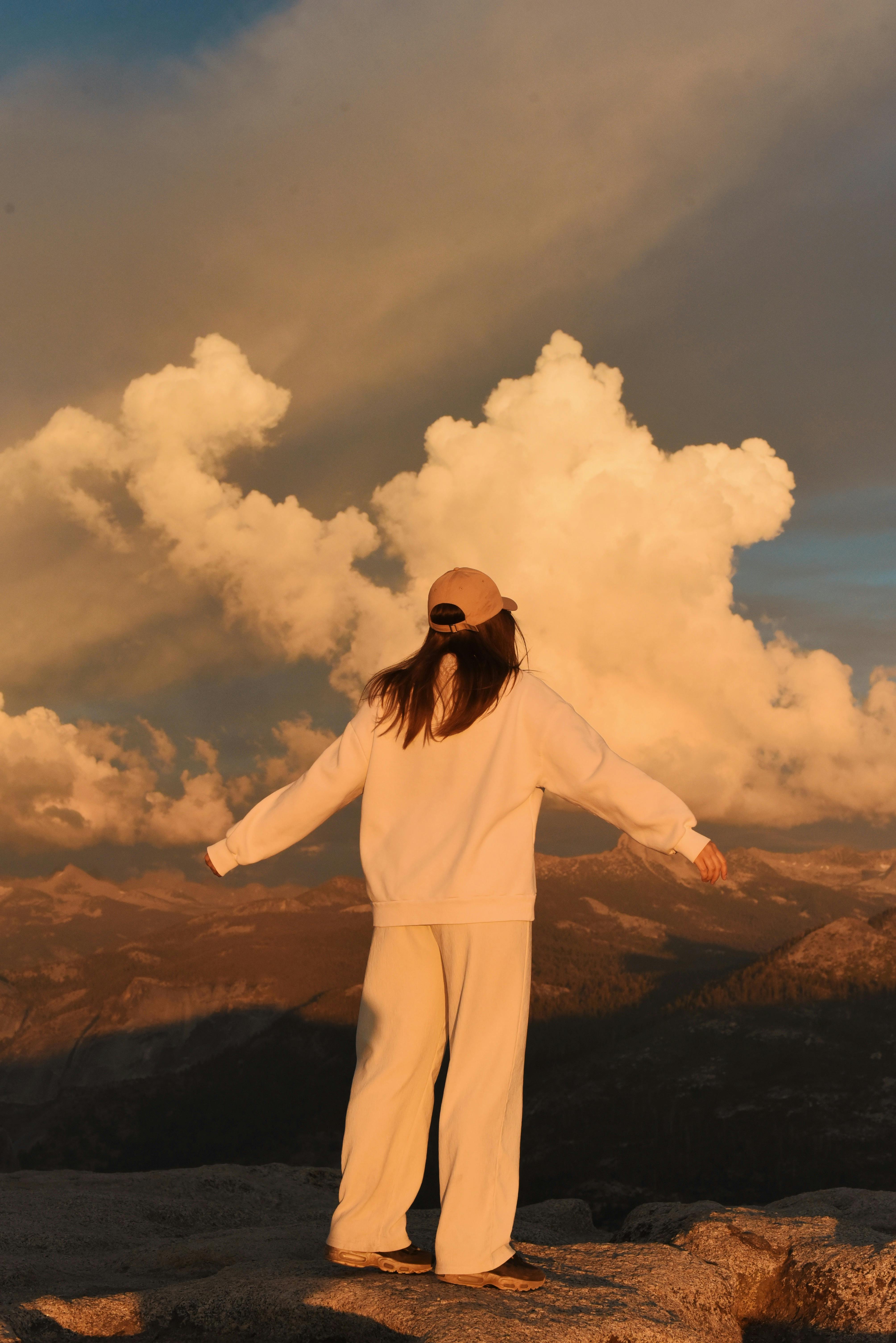 A person stands on a California mountain, arms outstretched, facing dramatic sunset clouds.