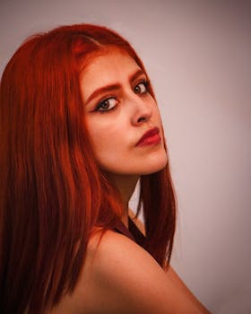 Striking portrait of a woman with red hair and intense eye makeup, set against a neutral background.