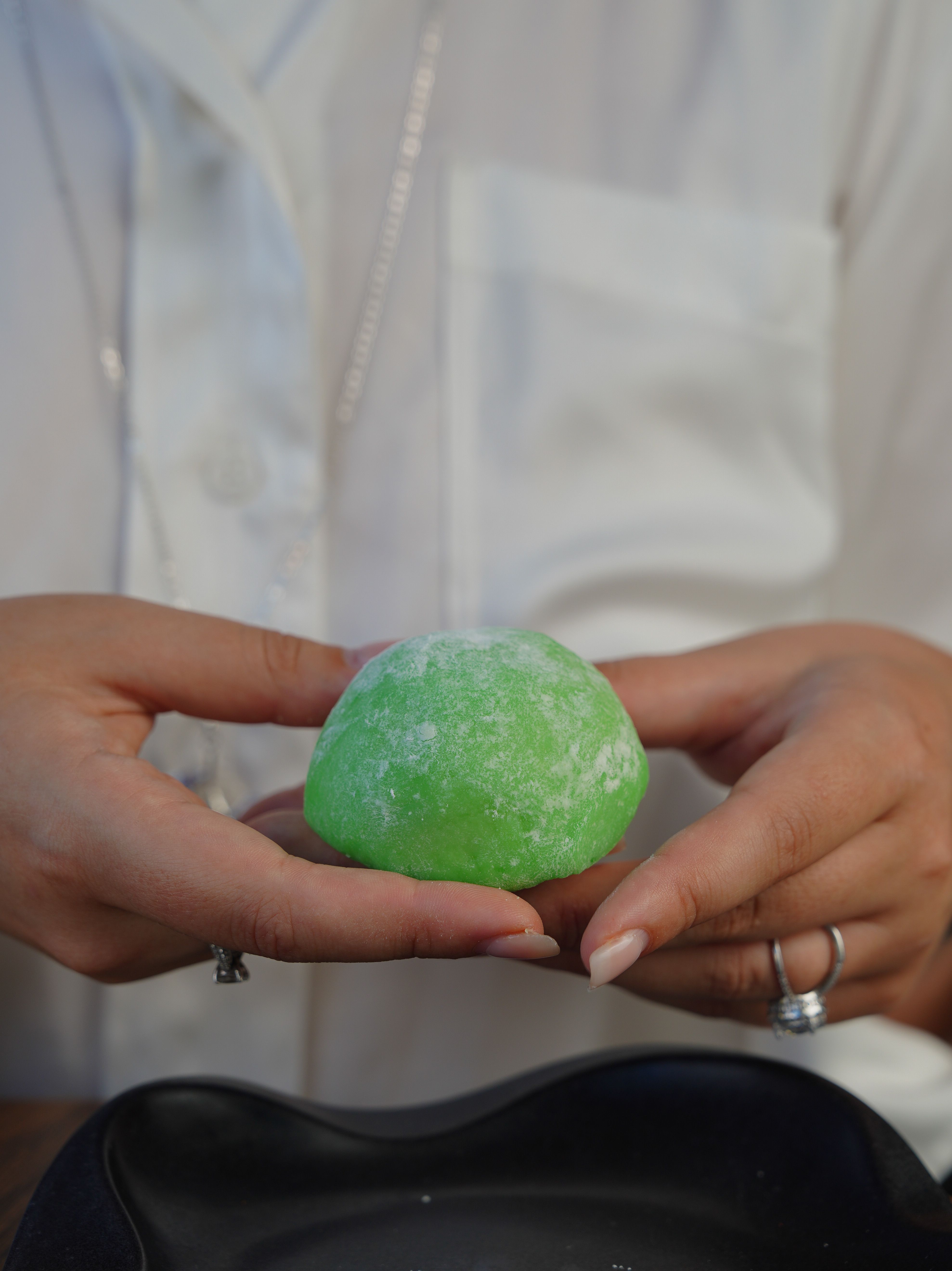 A close-up of hands gently holding a green mochi dessert over a black plate, indoors.