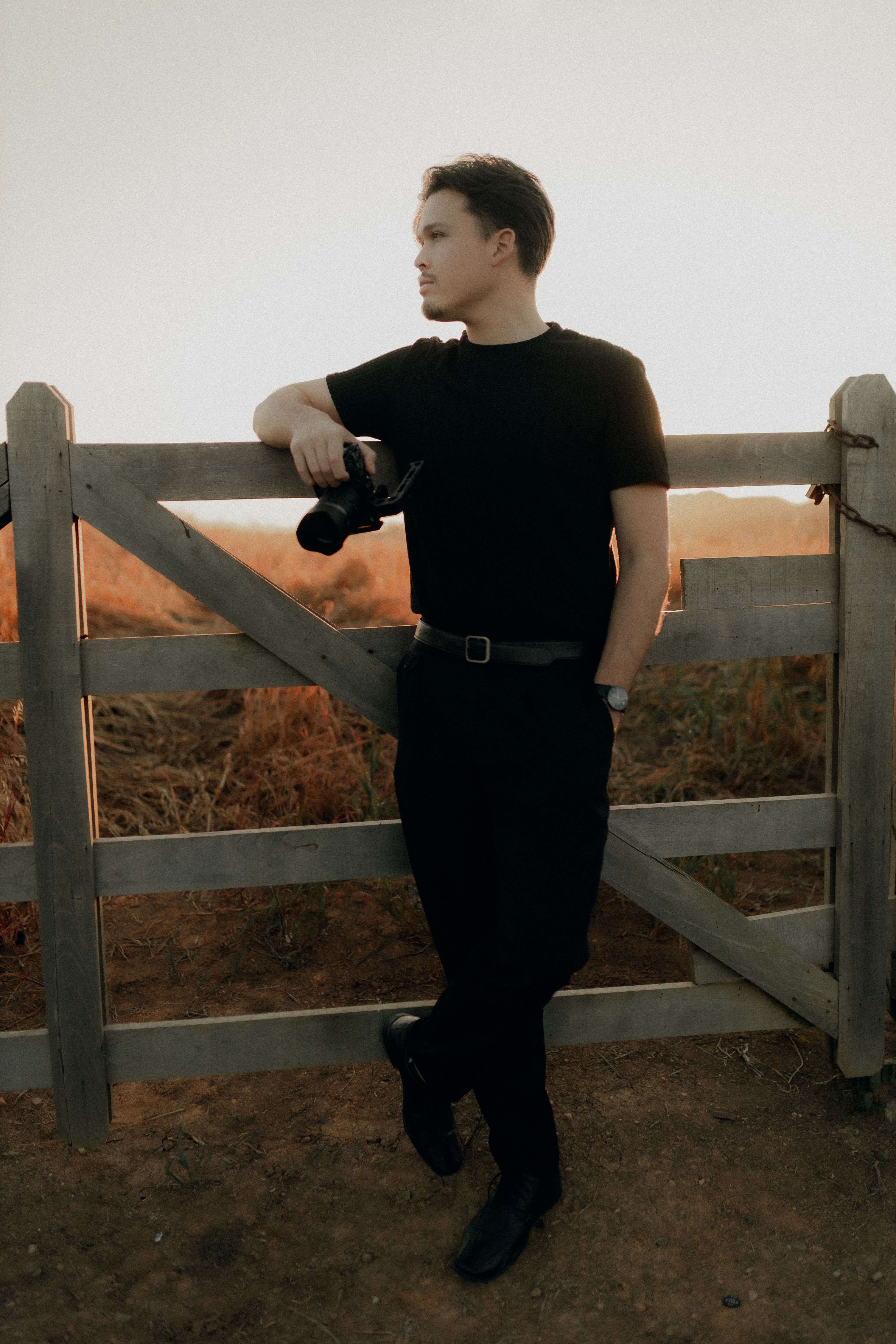 A photographer leans on a rustic gate with camera in hand, gazing at the sunset in a rural area.