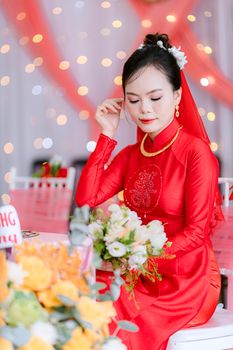 Bride in red dress with floral bouquet sitting at decorated wedding venue.