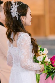 A bride in a lace gown holds a bouquet, captured in soft indoor lighting.