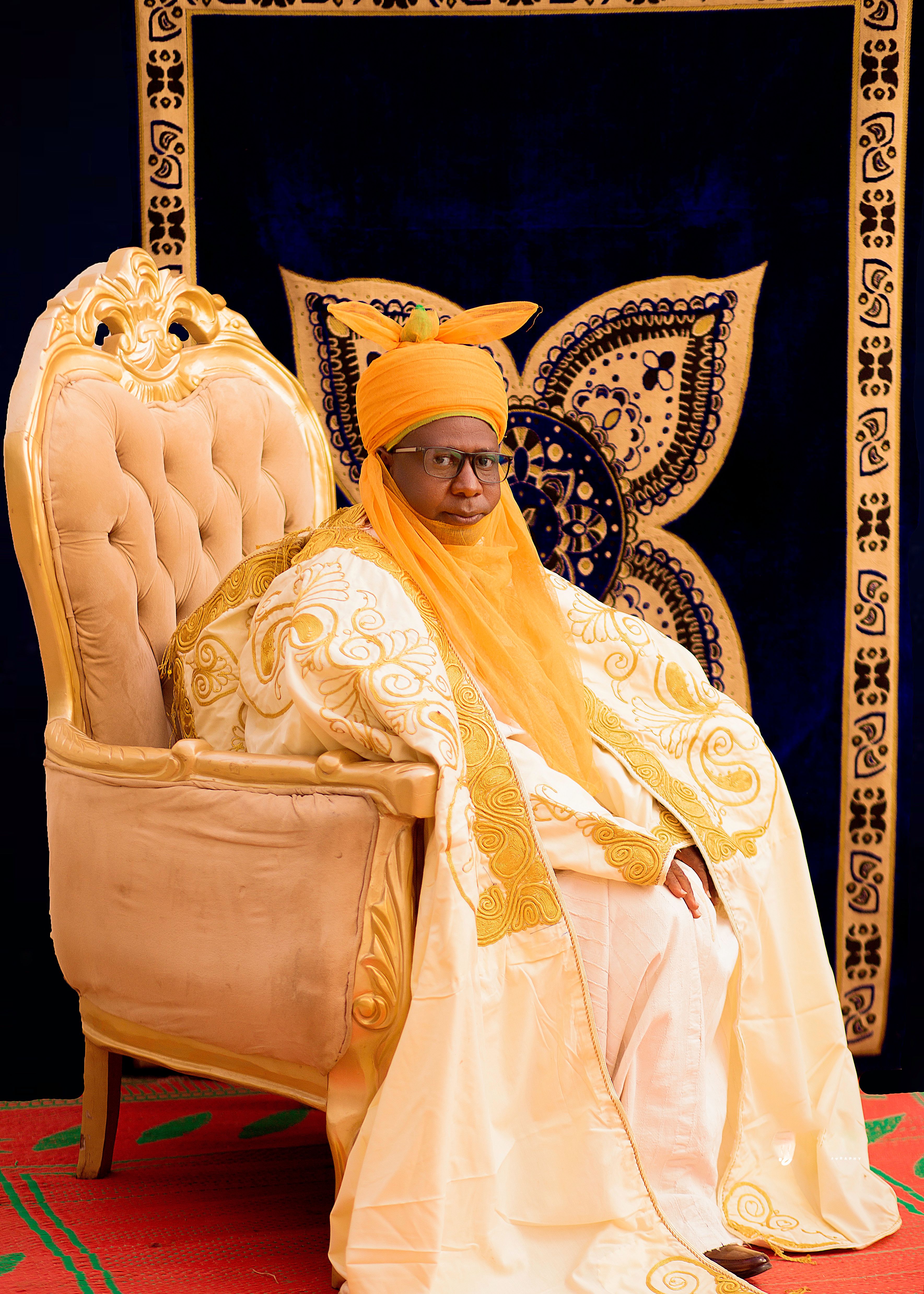 Man in ornate traditional attire seated on a decorative chair, exuding royalty.