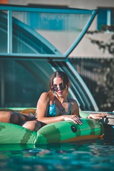 Stylish woman in sunglasses lounges on a green inflatable pool float, soaking in the sun.