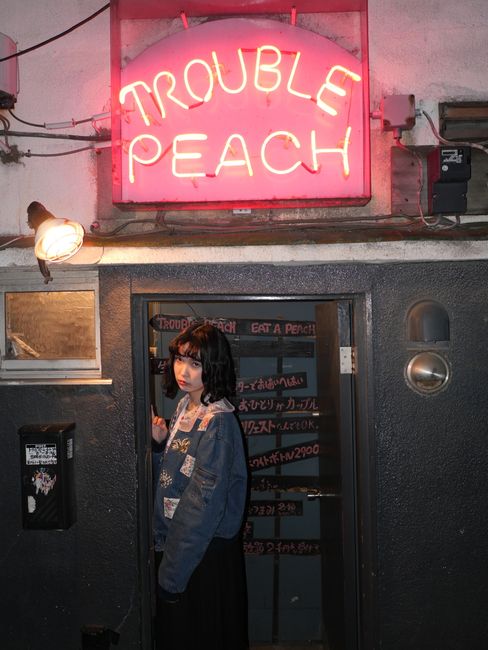 A woman stands under the neon-lit 'Trouble Peach' sign in Setagaya, Tokyo, capturing the vibrant nightlife.