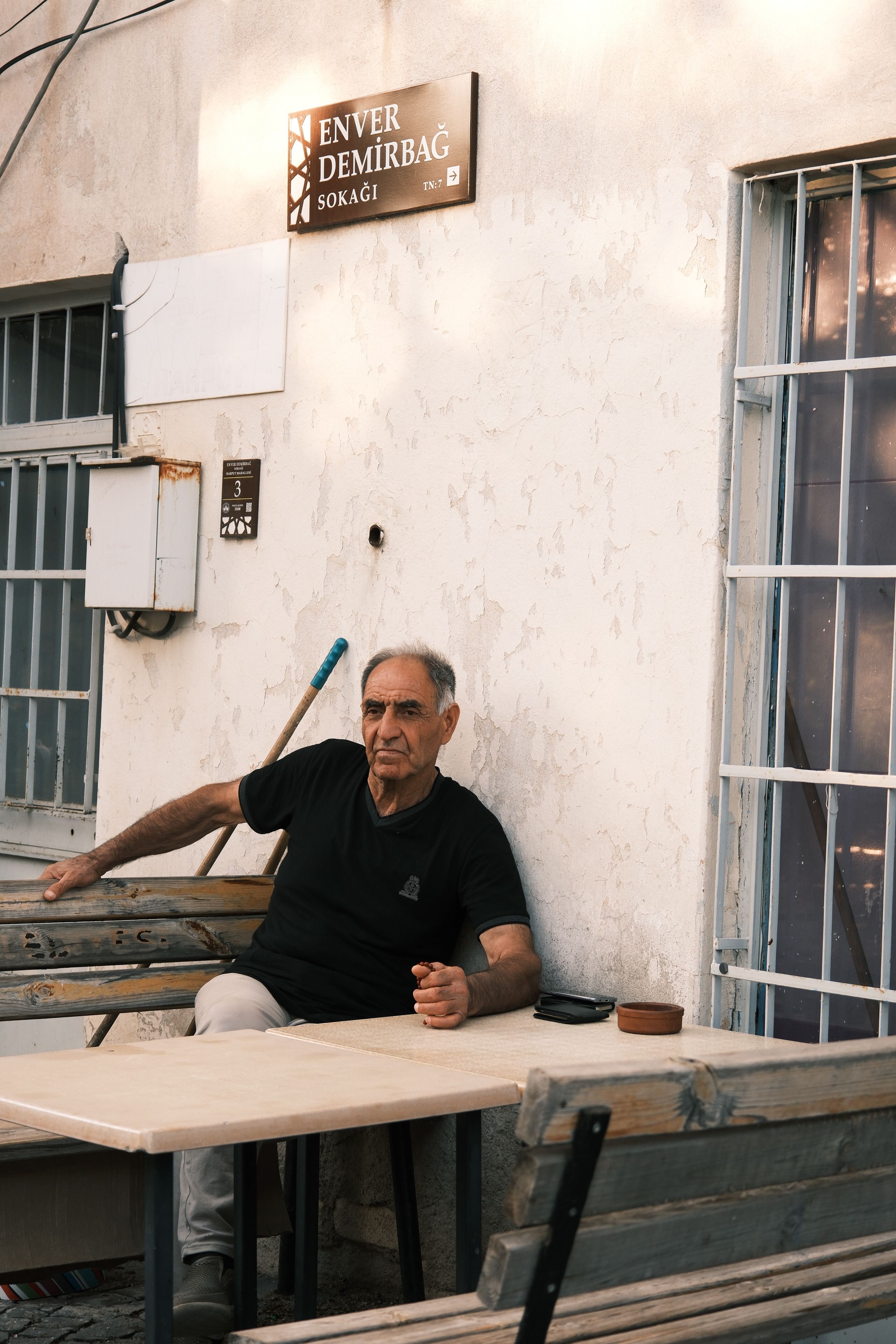 Elderly man sitting outside a rustic cafe on Enver Demirbağ Street, reflecting urban life.