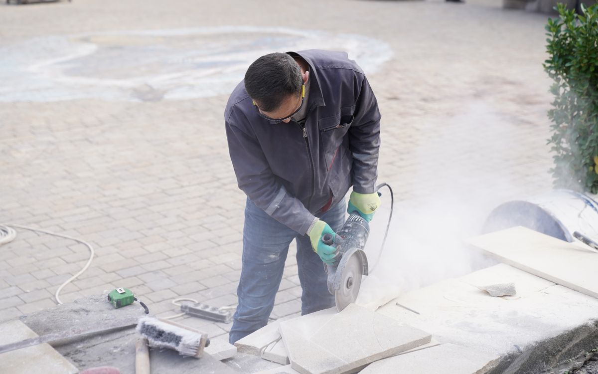 A construction worker uses a power saw to cut stone tiles outdoors, creating dust as he works.
