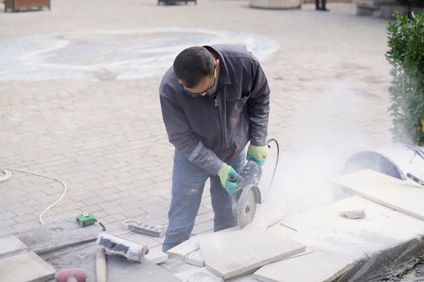 Worker wearing protective gloves uses an angle grinder to cut large stone or tile slabs outdoors, with dust and masonry tools nearby.