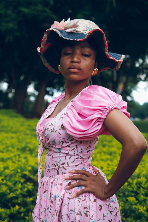 Free A stylish woman poses in a vibrant garden, wearing a pink floral dress and a wide-brimmed hat. Stock Photo