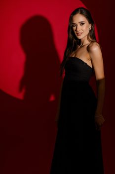 A woman in a stylish black dress poses against a red background with a striking shadow.
