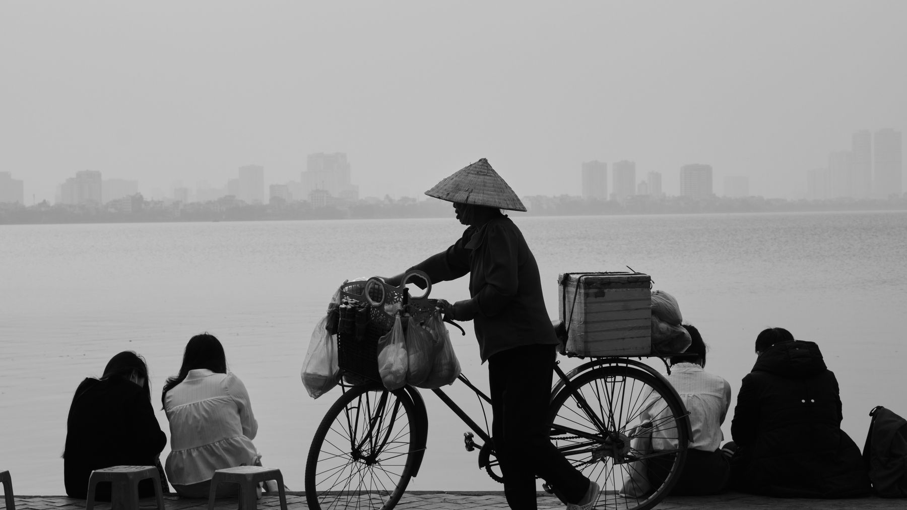 Black-and-white photo of a street vendor with a bicycle by Hanoi's lake, capturing daily life.