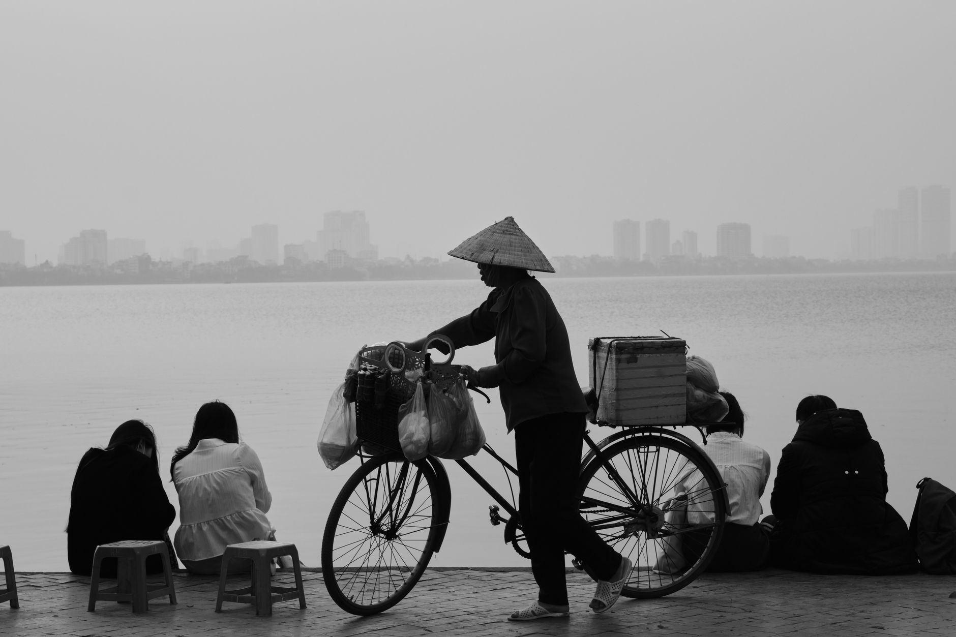 Black-and-white photo of a street vendor with a bicycle by Hanoi's lake, capturing daily life.