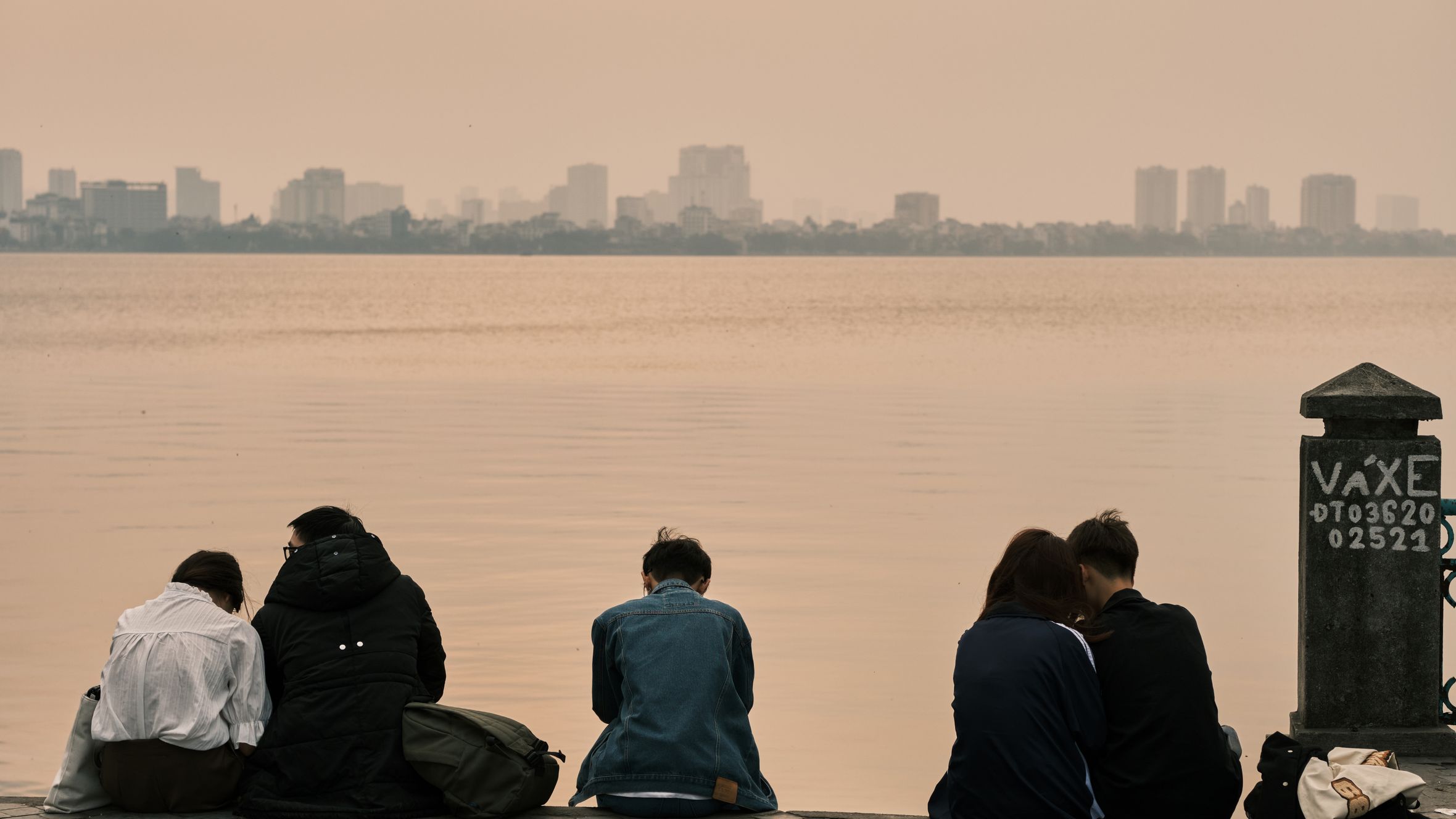 A group of young adults sitting by the tranquil West Lake in Hanoi, Vietnam during sunset.
