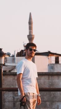 Young man in sunglasses stands confidently with an urban skyline and minaret in the background.