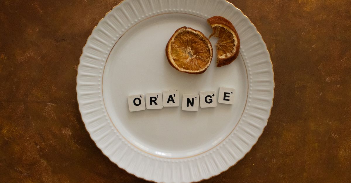 Artistic arrangement of dried orange slices and letter tiles spelling 'orange' on a white plate.
