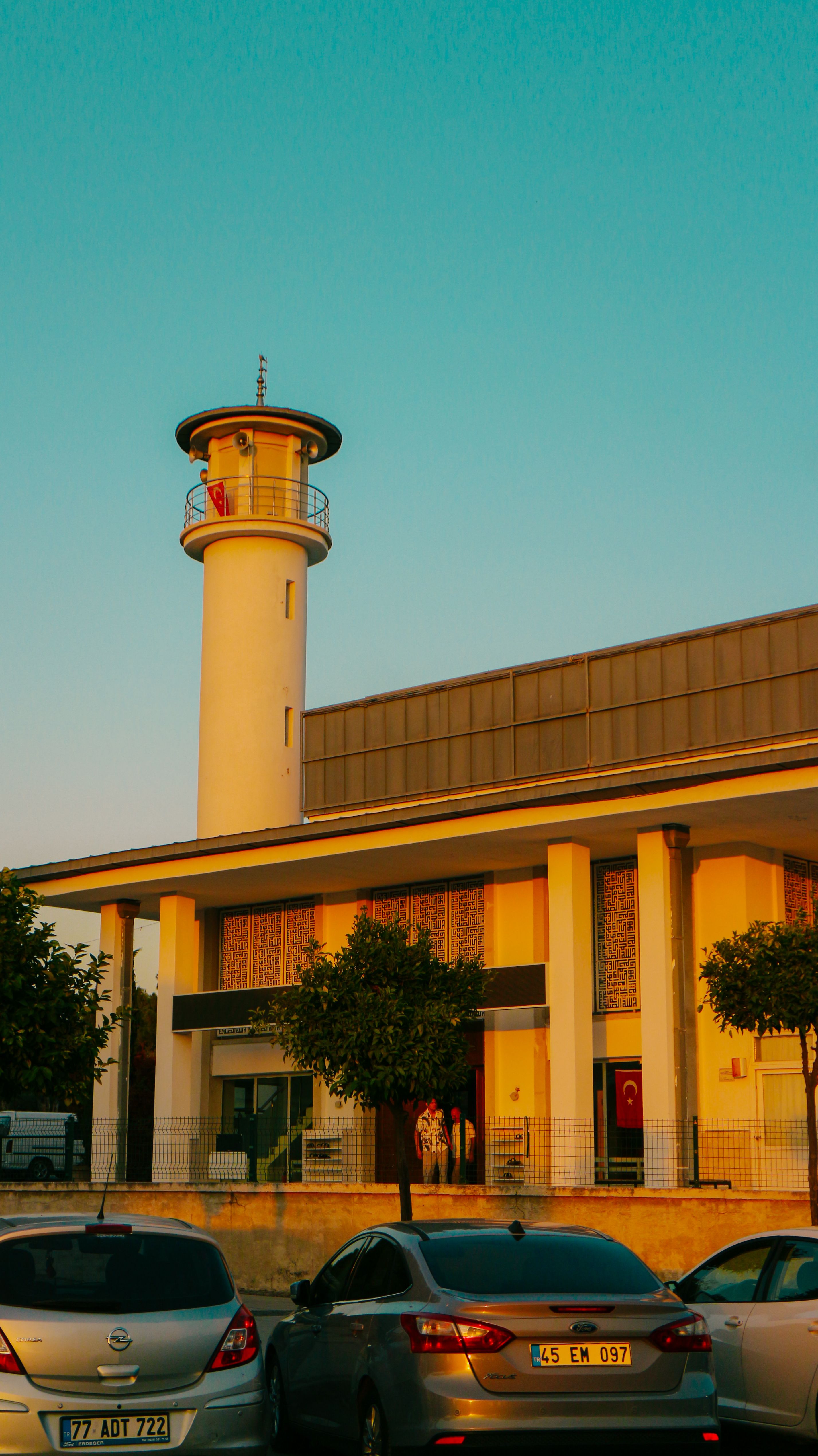 A contemporary building with a lighthouse tower illuminated by warm sunset light, capturing an urban scene with parked cars.