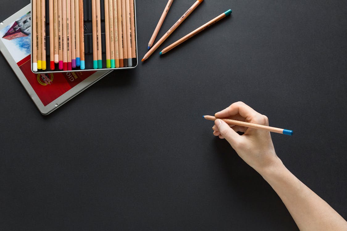 Uma mão segura um lápis sobre uma mesa escura com materiais de desenho
