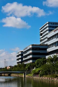 Modern office complexes beside a riverside, under a bright blue sky with clouds.