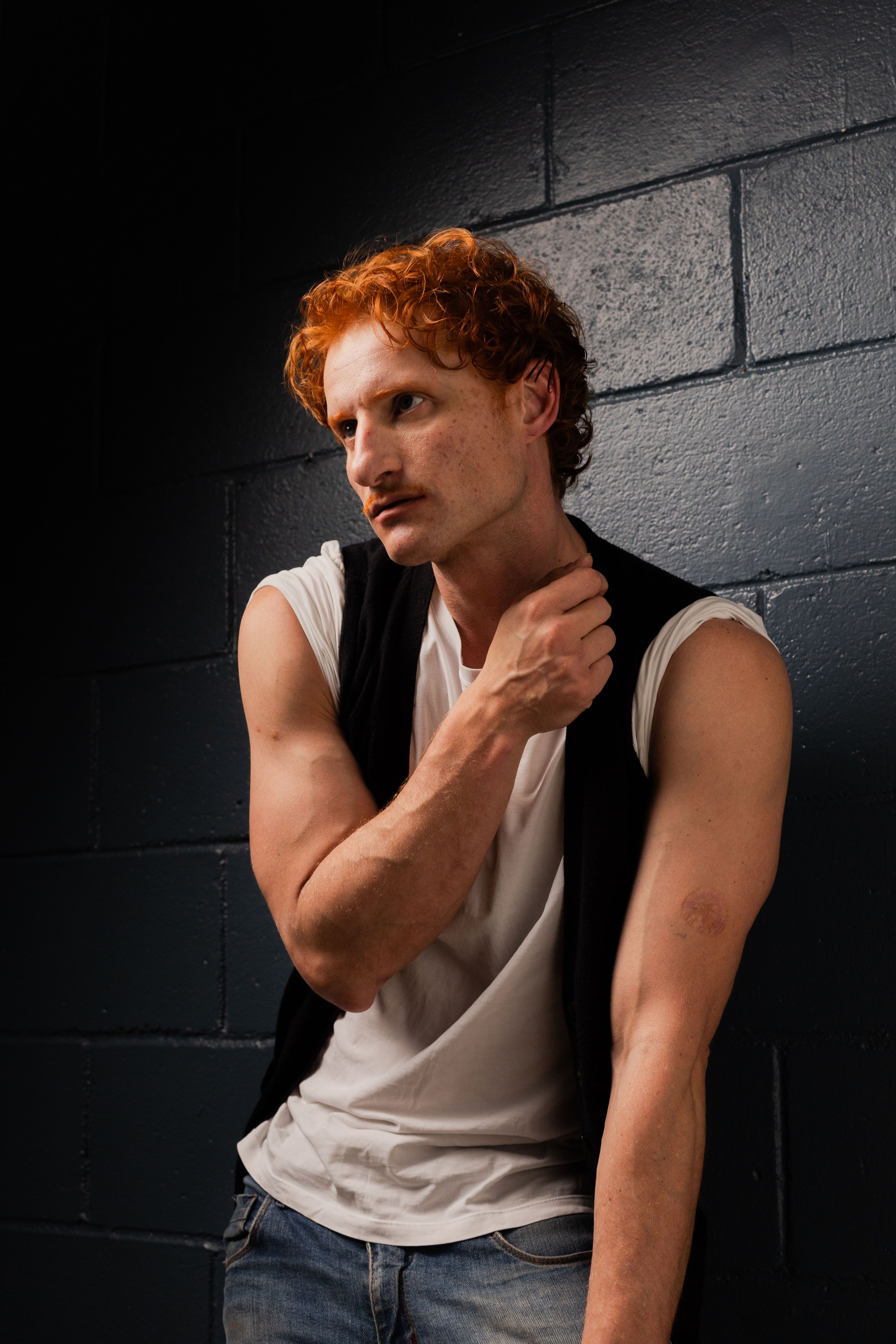 Portrait of a red-haired man in casual attire against a dark studio backdrop, exuding a pensive mood.