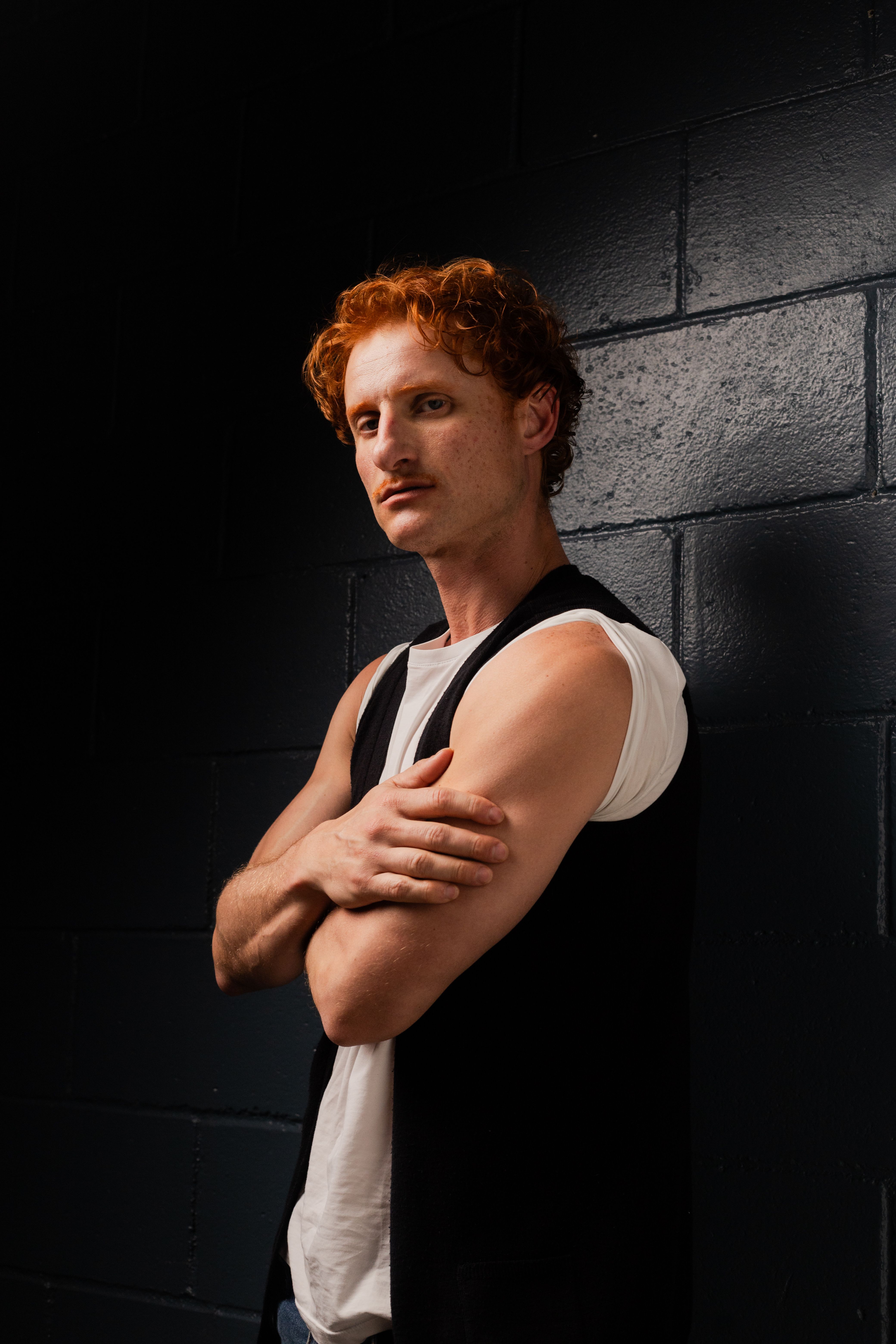 Red-haired man posing against a dark wall with moody lighting in a studio setting.