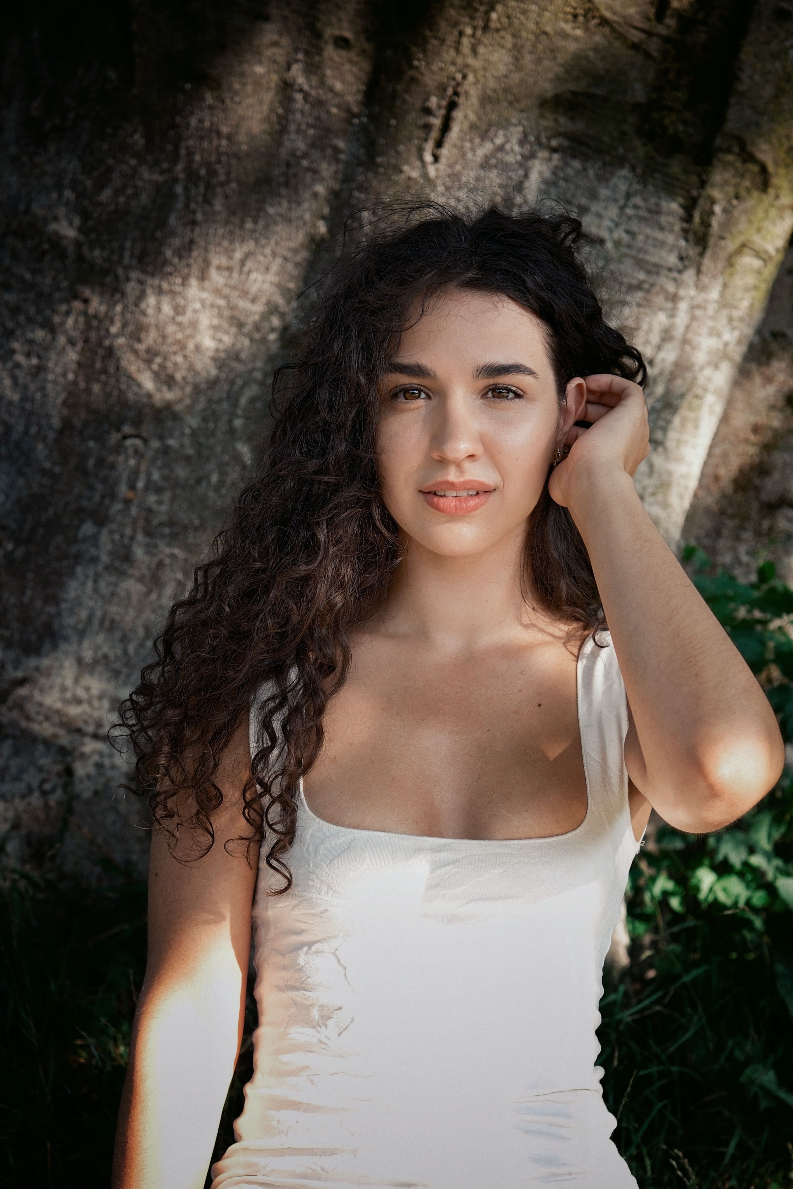 Portrait of a young woman with curly hair in a white dress sitting under a tree.