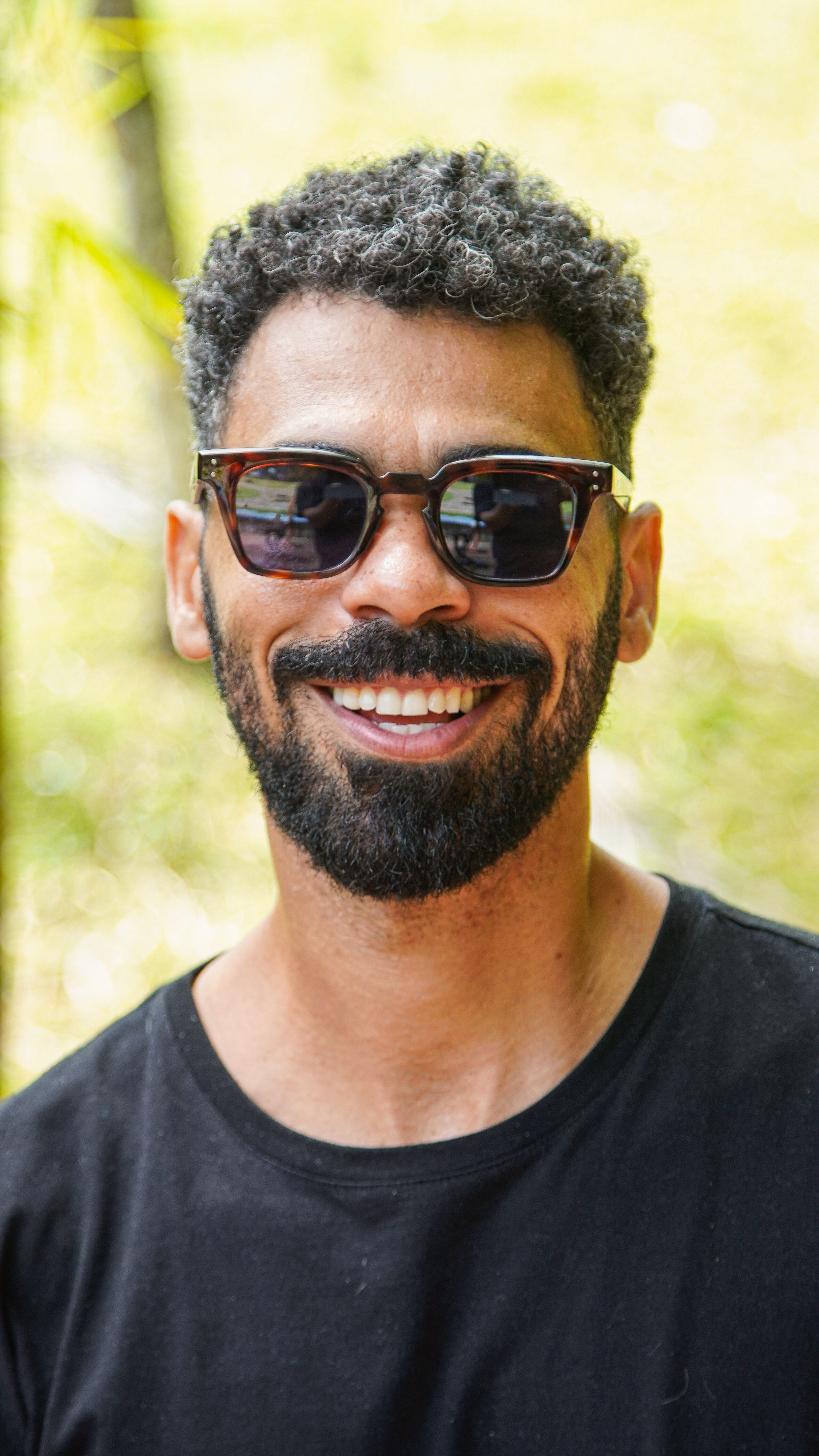 A cheerful adult male smiling confidently while wearing sunglasses in an outdoor setting.