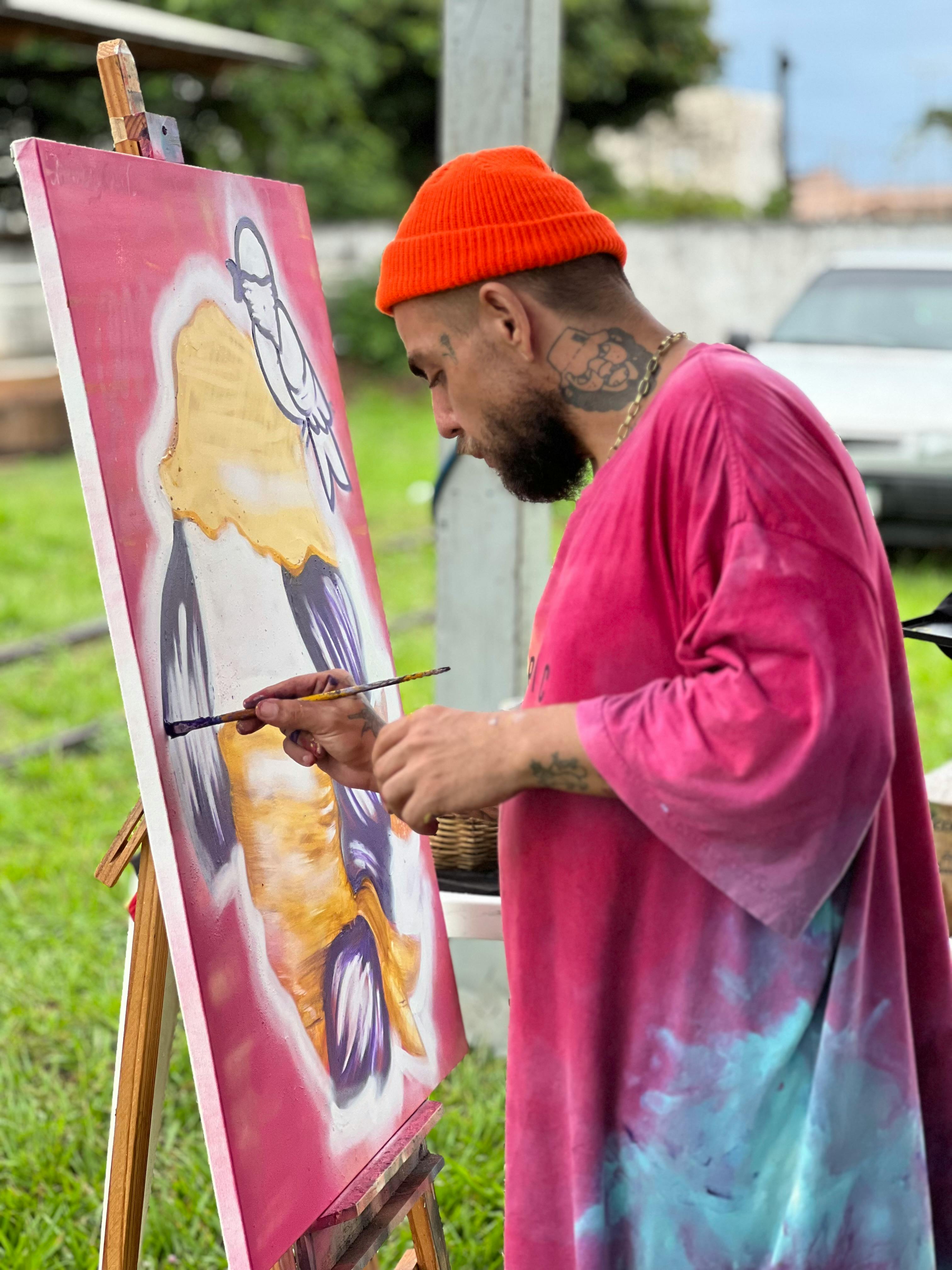 Artist in orange beanie painting on canvas outside with grass background.