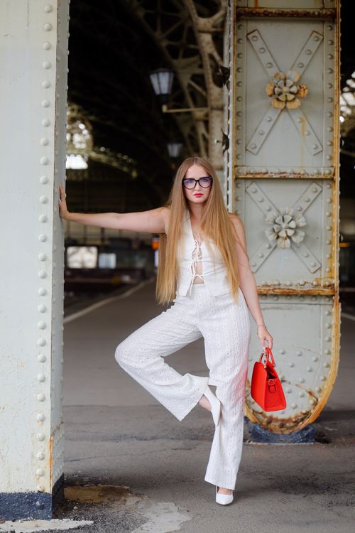 Free Fashionable woman in white outfit poses with red bag in industrial urban setting. Stock Photo