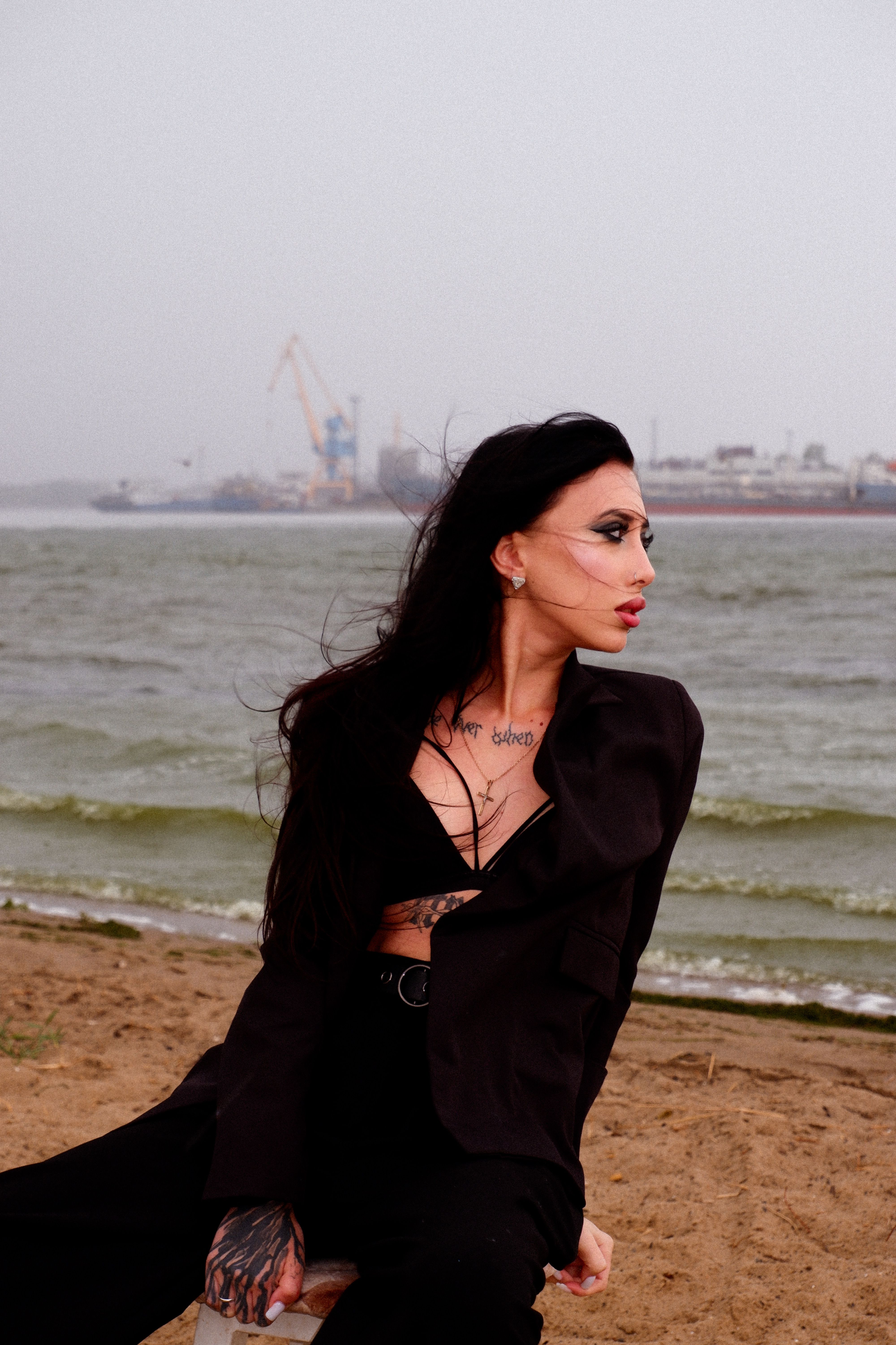 Elegant woman in black on beach with industrial port in background.