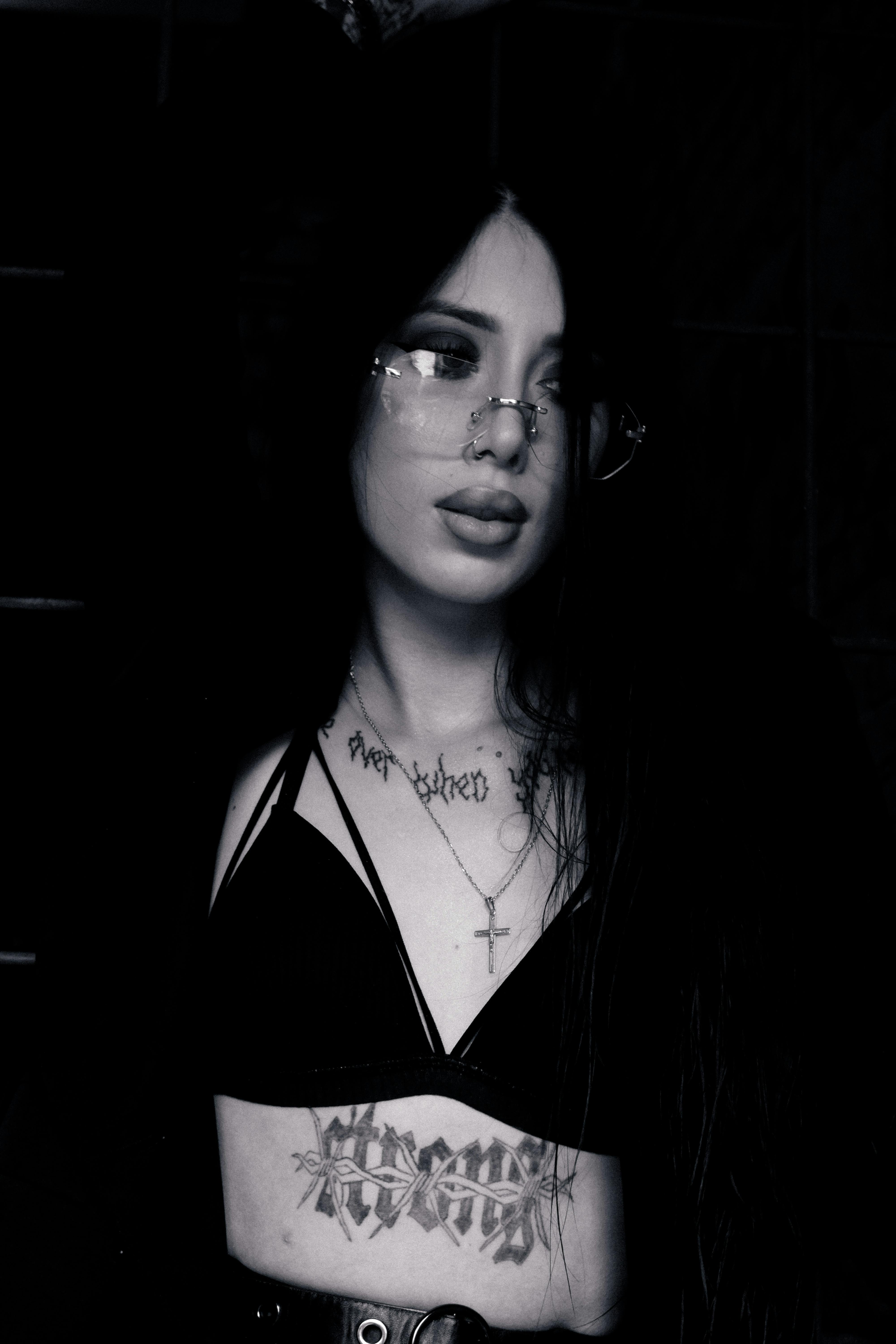 Moody black and white portrait of a tattooed woman wearing glasses and a cross necklace.