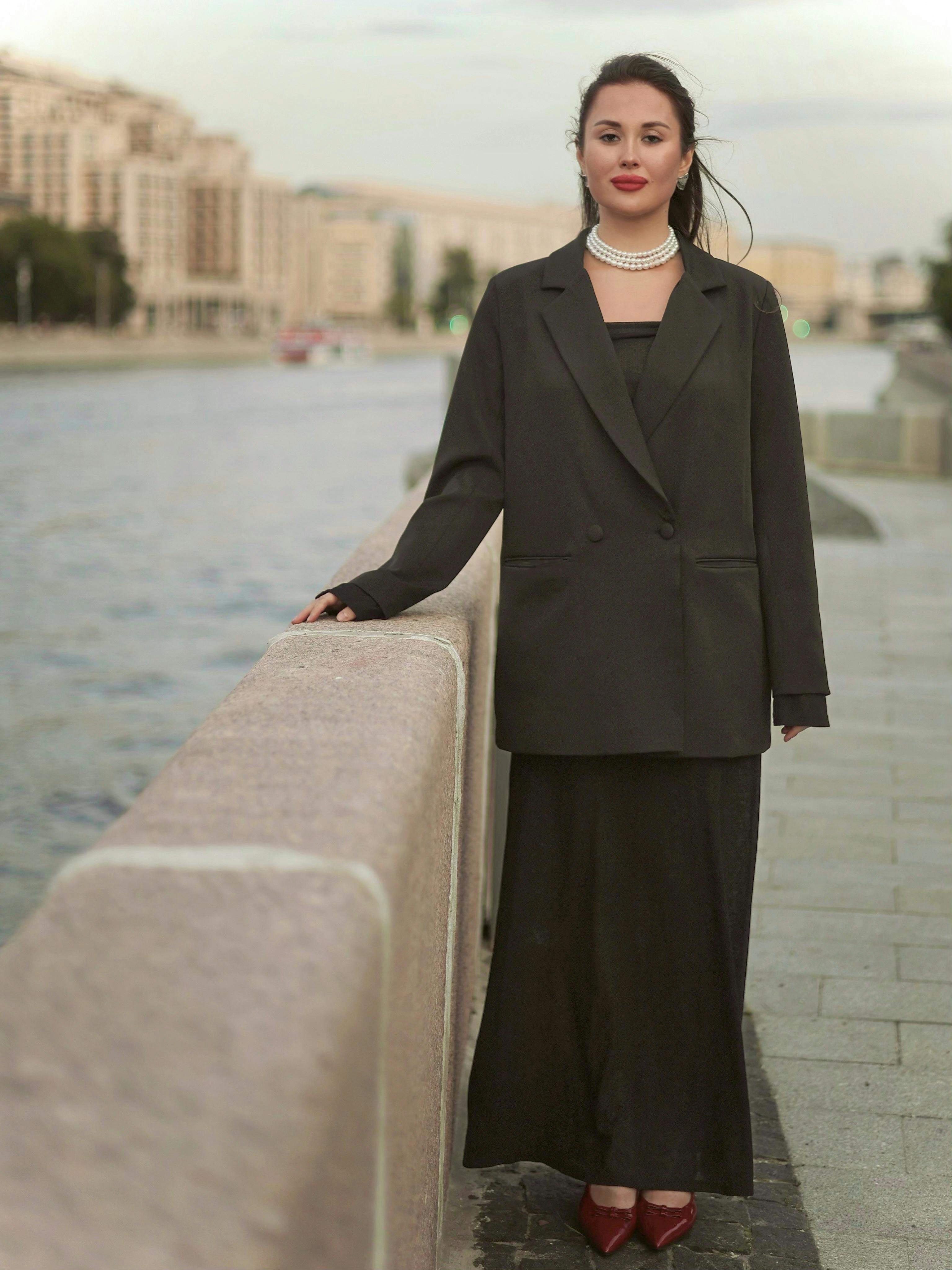 Young woman in a black coat stands by a riverside, exuding elegance.