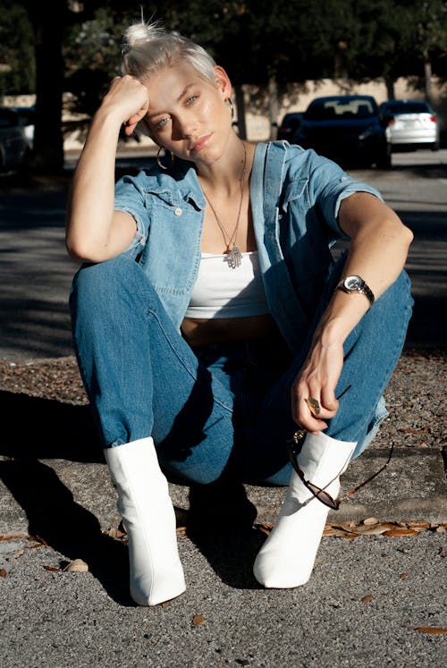 Free Full body of modern woman with short blonde hair wearing denim outfit with trendy white boots sitting on sidewalk looking at camera Stock Photo