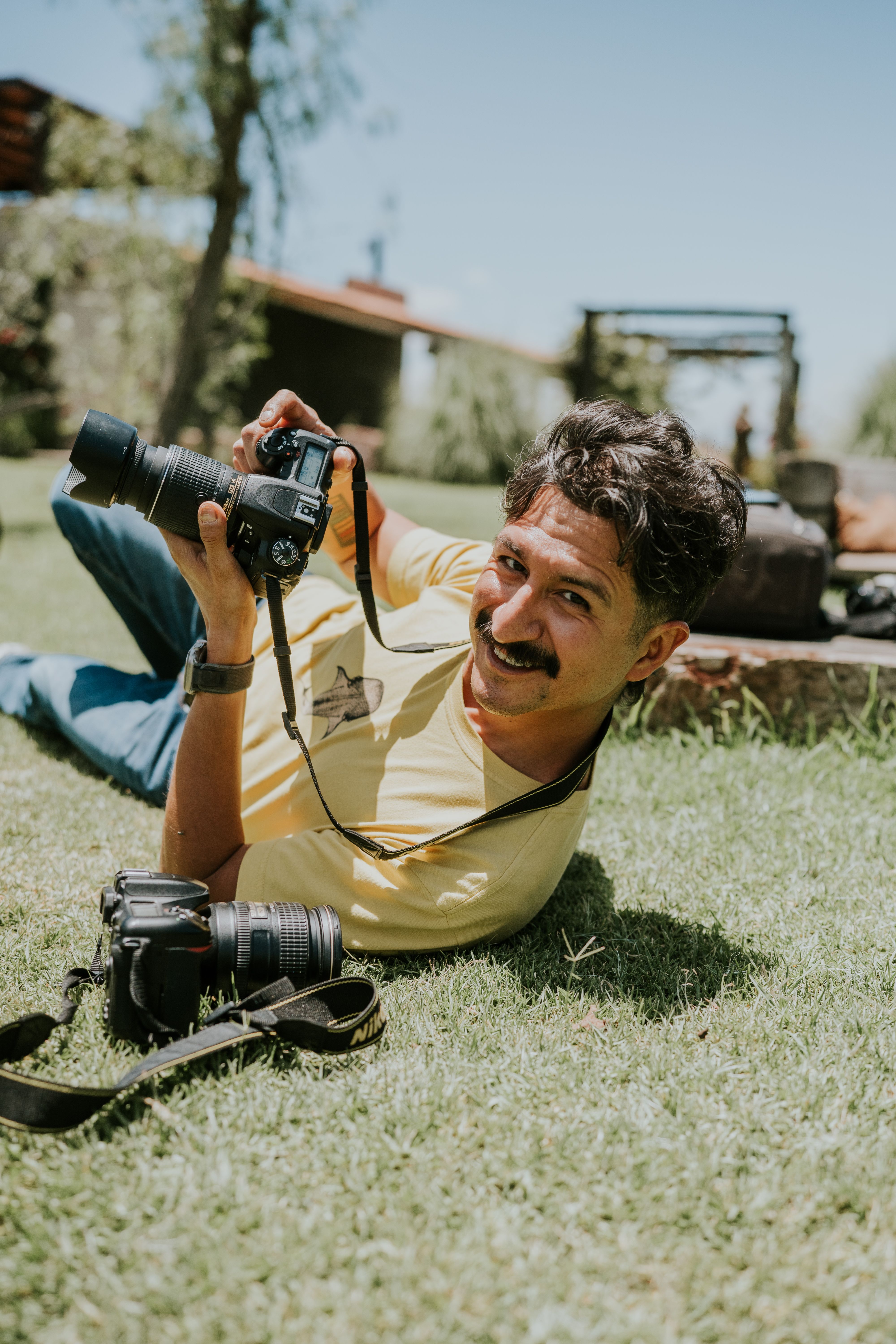 Smiling man with a camera lying on the grass in a sunny outdoor setting, showcasing photography passion.