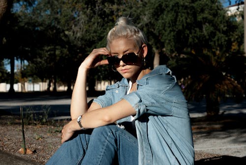 Free Serious young woman with hair bun wearing denim coat and pants looking at camera over black sunglasses while lounging in square in sunlight Stock Photo