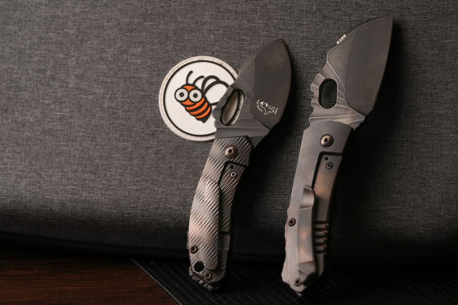 Two tactical knives resting on dark fabric with a bee patch, highlighting knife design details.