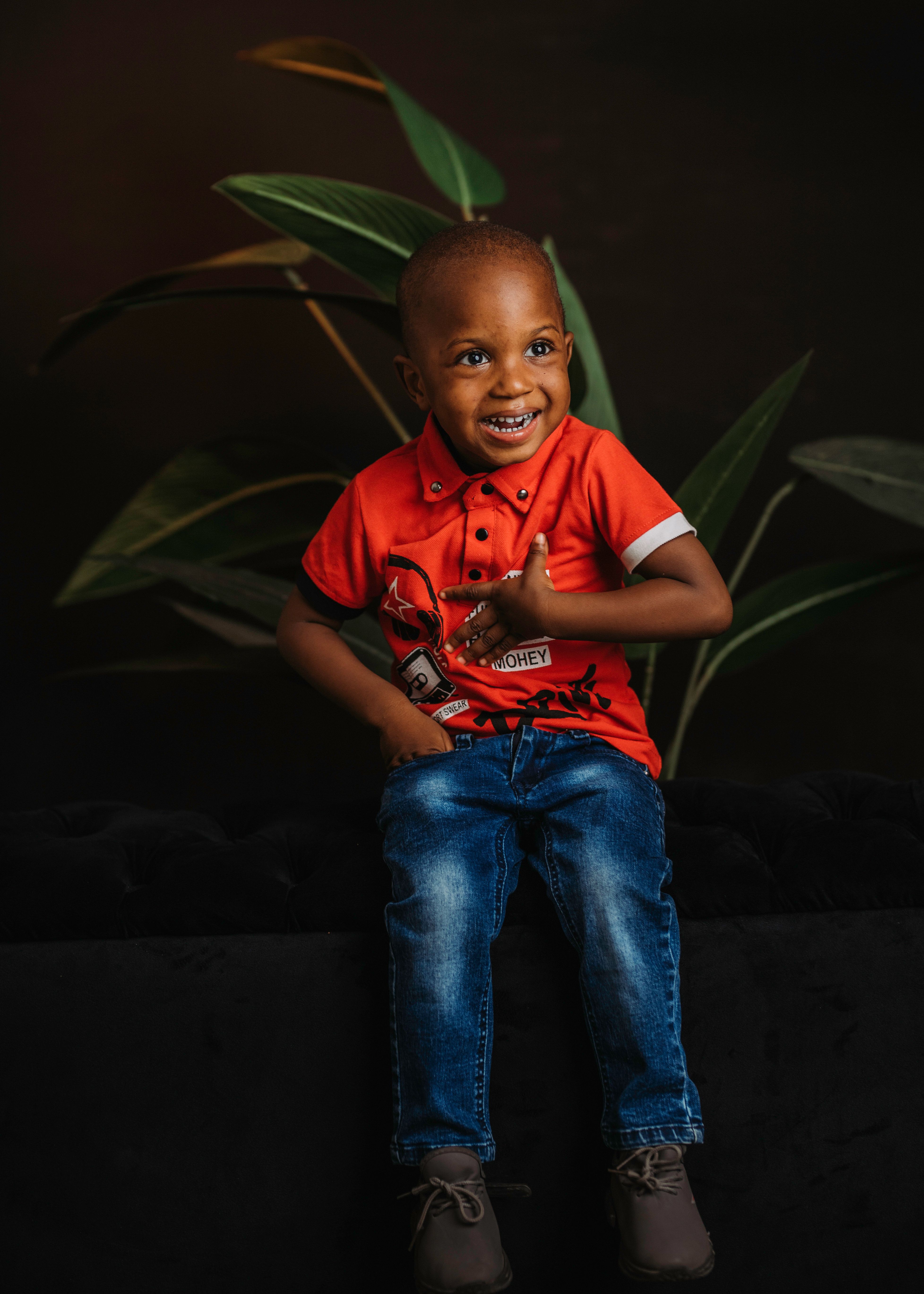 Delightful toddler in a red shirt, sitting joyfully indoors with plant leaves.