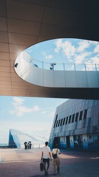 A couple walks under a modern architectural structure with a circular sky opening, showcasing urban design.